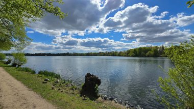 Bulutlu bir gökyüzünün altında yemyeşil ağaçlar ve çimenler arasında uzanan sakin bir göl bulutlu gökyüzünü yansıtan suyla sakin bir doğal manzara yaratıyor.