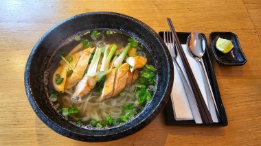 Bir kase Pho tavuklu şehriye çorbası, ahşap bir masanın üzerinde oturup, konforlu bir tabak oluşturmak için pişirilmiş malzemelerin ve sıvıların karışımını sergiliyor.