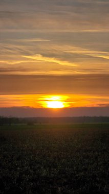 Kehribar güneşi doğal bir manzaranın üzerinde batıyor, uzaktaki tarlaya sıcak bir ışık yayıyor, bulutlar gökyüzünü kırmızı ve turuncu Wunstorf Hanover Almanya renkleriyle boyuyor.