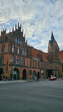 Bulutlu gökyüzüne karşı kulesi olan büyük bir tuğla bina ayakta duruyor. Hanover Almanya 'sının aşağısındaki asfalt yol yüzeyinde araçlar geçerken pencereler ön cephesini kapatıyor.