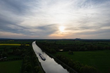 Gün batımında bir nehrin manzarası. Parıldayan suda süzülen bir tekne, yemyeşil düzlüklerle çevrili ve kargo gemisi mavna ile dolu çarpıcı bir gökyüzü.