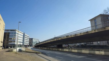 Arka planda gökyüzüne karşı binalar bulunan yol üzerinde bir köprü barındıran bir şehir tasarımı. Asfalt yol yüzeyi bitkiler ve modern Almanya mühendisliği ile tamamlanmıştır.