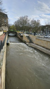 Köprünün altından geçen ve kıyıları kaplayan ağaçlarla dolu pitoresk bir nehir, binaların ve asfalt yolların kentsel manzarasına zıt düşüyor.