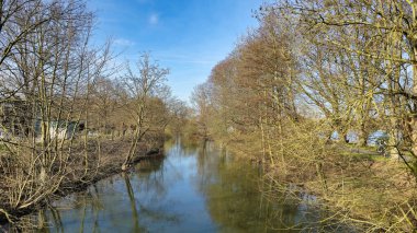Sakin bir nehir, güneşli bir gökyüzünün altındaki ağaçların arasından akar ve doğal güzelliği resmeden Leine Maschsee Hanover 'ı yaratır.