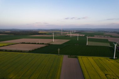 Rüzgâr türbinlerinin uçsuz bucaksız bir çayıra dağılmış nefes kesici hava manzarası bulutlu bir gökyüzü altında, ufukta ağaçlar ve yollar var 5K insansız hava aracı Hanover Wunstorf Kolenfeld Almanya 