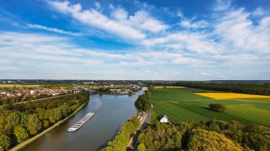 Bereketli tarlalar ve ormanlar arasında süzülen sarmal bir nehrin, tüylü beyaz bulutlarla dolu açık mavi bir gökyüzü altında, güzel bir doğal manzara sergiliyor. Mittellandkanal