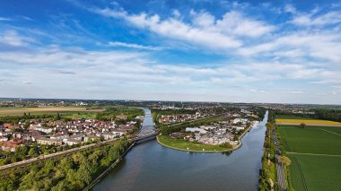 Ağaçların, tarlaların ve kentsel binaların ortasından geçen bir nehrin havadan görünüşü. Su kaynakları yeşillik ve yukarıdaki bulutlu gökyüzü ile tezat oluşturuyor Mittellandkanal Hanover Seelze