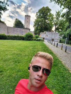 Güneş gözlüklü ve kırmızı gömlekli bir adam arka planda ağaçlarla dolu bulutlu bir gökyüzünün altındaki yeşil bir parkta selfie çekiyor.