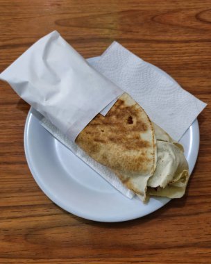 Basit beyaz bir tabakta, peçeteyle servis edilen bir tortilla dürümü. Bu fast food yemeği temel malzemeler olarak taze ürünler ve pişmiş ürünler içerir.
