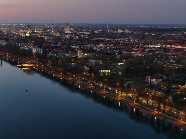 Gün batımında nehir kenarındaki canlı şehir manzarası, Maschsee Hannover binalarının parlayan ışıklarını vurguluyor.
