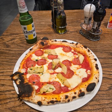 Lezzetli pepperonili ve enginarlı lezzetli bir pizza. Köhne ahşap bir masada soğuk bir içecekle servis ediliyor.