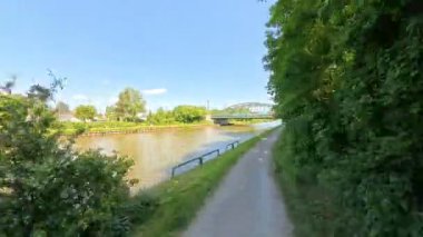 Köprülerin altından süzülen, yemyeşil ağaçlar ve açık mavi gökyüzü ile çevrili nehir kenarındaki manzaralı bir yolun tadını çıkar.