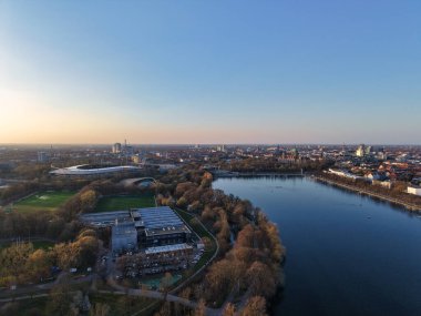 Şok edici hava manzarası, gün batımında nehir kenarında canlı bir şehir yakalıyor, parkları ve mimariyi gözler önüne seriyor. Maschsee Hannover