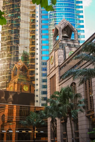 An old sandstone building with a bell tower is reflected in the windows of a glass skyscraper. Old colonial architecture and modern style side by side. City center, Hong Kong.