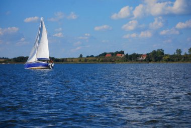 Gölde mavi bir yelkenli yelken açıyor. Kıyı ve arka planda bazı evler. Masuria 'da yat gezisi (Mazury). Masurian göl bölgesi, Polonya.