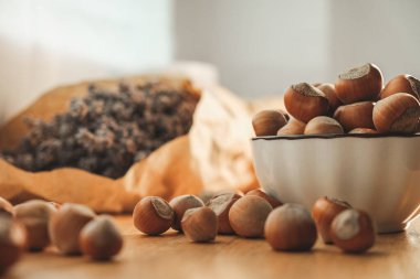 Bir kase taze fındık, dağınık fındıklarla çevrili ahşap bir masanın üzerinde duruyor. Arkasında, kurumuş çiçek torbaları davetkar mutfak ortamını sıcak tutar..