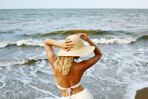 Blonde Woman in Fashion Summer Style Standing at Sea and Holding Hat. Luxury Lifestyle Rear View. Summer Vacation Concept with Ocean. Soul Relaxing, Spirit Calm