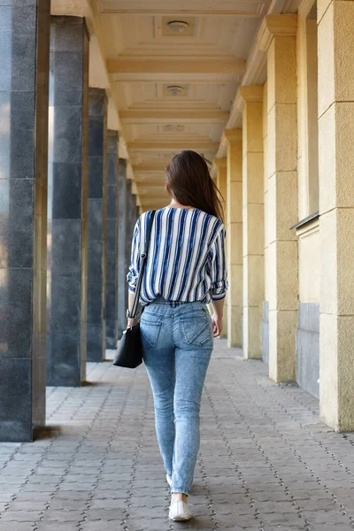 Rear Behind View Photo of Charming Lady Walking around the City through the Colonnade. Beautiful free Woman in Modern Clothes, Denim Skinny Jeans and Blue Blouse