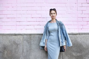 Kot ceket ve gri elbise giyen şık bir kadın manken. Pembe duvar arkaplanı olan açık hava portresi. Sokak tarzı, moda tarzı ve modaya uygun kentsel görünüş. Pembe Duvardaki Kadın Sokak Tarzı Çekimi. Swag Girl kot ceket giyiyor, gri elbise giyiyor.