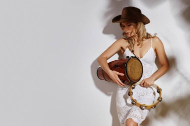 Beyaz örgü dantelli, kovboy şapkalı, davul ve tef tutan boho tarzı bir kadın. Bitki gölgeleriyle süslenmiş beyaz bir duvara karşı durarak kaygısız yaz titreşimleri yayıyor. Kompozisyon bohem modasını, müzikali harmanlıyor.