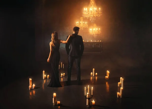 gothic couple stands in dark room woman hugs man in business suit. black elegant dress. ball carnival party, luxurious scenery many candles burning. Fantasy girl vampire queen guy king back rear view