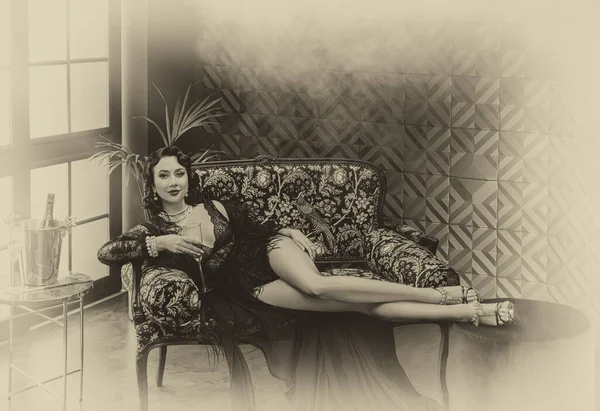1920年代の古いスタイルのセクシーなフラッパーの女性はソファ シルクのペノギアドレスダチョウの羽にあります シャンパンを持ってる女の子 豪華な室内 黒と白のセピアヴィンテージ写真 — ストック写真