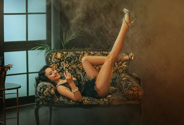 20世纪20年代风格快乐的女人躺在沙发上 时尚模特穿着光秃秃性感的长腿高跟鞋和黑色短裙 复古的女孩手拿着话筒 经典的室内浓烟 — 图库照片