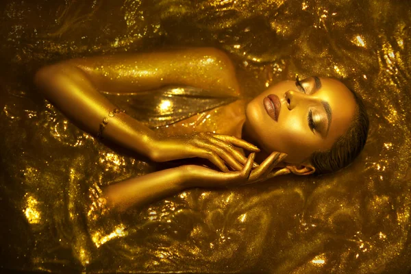 Portrait Closeup Beauty fantasy woman face in gold paint. Golden shiny skin. Glamorous fashion model girl image goddess lies bathed in liquid gold. Professional metallic glowing luxury royal makeup.