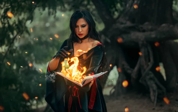 ファンタジーハロウィン女性の魔女は手に古い燃焼魔法の本を保持し 明るい炎の火の光の中でスペルペーパーページを読み取ります 灰の中のゴシックガールセクシーな顔 黒いドレスの衣装 暗い夜の森の木 — ストック写真