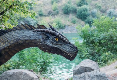 Fantasy art portrait real dragon head close-up sharp teeth beautiful eyes muzzle in dark gray-green scales, spikes. Creative big dinosaur toy of Jurassic period. Background summer nature green trees.