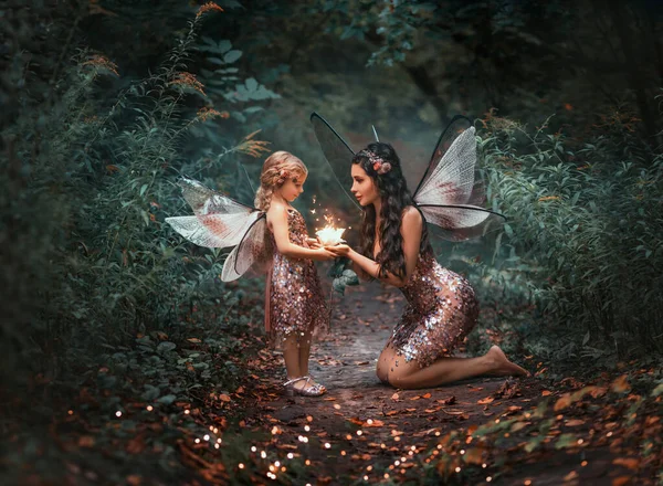ファンタジー妖精の女性のお母さんは小さな幸せなピクシーの女の子に魔法の光輝く花を与えます 暗い夜夏の緑の森の木 蝶の衣装ピンクのドレス おとぎ話の世界で子供の笑顔 — ストック写真