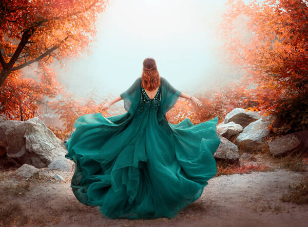 Fantasy woman queen red hair runs in autumn forest. Girl in long elegant royal evening vintage green dress. silk fabric skirt flying fluttering in wind. Orange tree. Art photo bare open back, no face