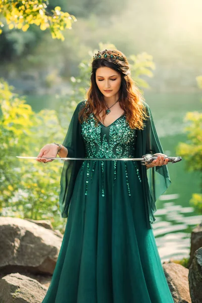 Fantasy warlike red-haired woman queen holding silver sword in hands praying before battle. Ritsa ritual of sacrifice. Green silk dress long elven sleeves, golden crown emeralds gems medieval style.
