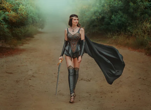 Fantasy woman viking princess warrior walks down road. Warlike girl nordic style sexy valkyrie goddess holding sword in hand. Black Leather costume armor cloak flies in wind. Fog green nature summer