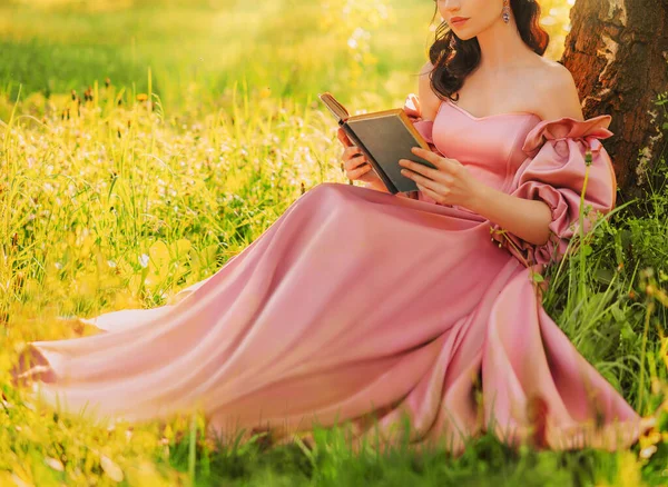 幻想中的女人坐在树下 手里拿着浪漫的书 看着小说般的笑脸 嘴角紧闭 粉色长裙 仙女在夏天的自然绿草魔法阳光下 艺术摄影真人 — 图库照片