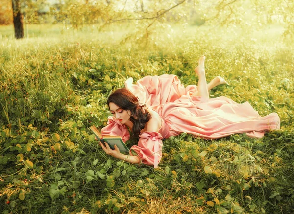 ファンタジー幸せな女性の森の芝生の緑の芝生の上にある手の魔法の本の小説の古いスタイルで 明るい魔法の太陽の光を保持します 長いピンクのヴィンテージのドレスでロマンチックなかわいいセクシーな女の子 中世の女性魔術師 — ストック写真