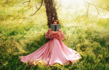 Fantezi kadın elinde romantik bir kitapla ağacın altında oturmuş roman okuyor. Pembe, uzun, eski bir elbise. Bahçedeki peri prenses yaz doğası yeşil çimenler güneş ışığı saçıyor. sanat fotoğrafı gerçek insanlar