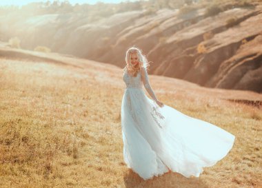 Mutlu, güzel, neşeli gelin dans ediyor, dans ediyor, eğleniyor, uzun, gri, açık renkli gelinlik rüzgar esintisiyle sallanıyor. Kız sarı saçlı, gülen surat. Sonbahar doğası ilahi sihirli güneş ışığı