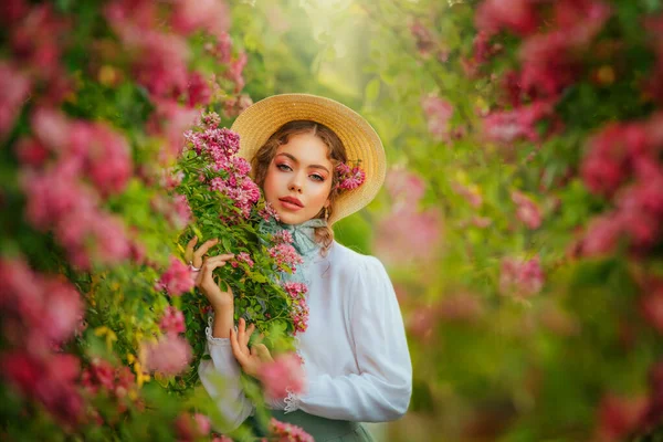 Art Fantasy young woman in vintage straw hat looks at camera blue eyes, girl princess pretty face red pink lips make-up, vintage old style white dress, garden green tree, rose flowers summer nature.
