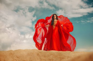 Arap seksi kadın kum tepelerinde mavi gökyüzünde duruyor. Yaratıcı tasarım giysileri kırmızı uzun elbise kumaş tren sineği rüzgarda sallanıyor. Doğulu kız manken pozu, güzel bayan yüzü siyah kıvırcık saçlı.
