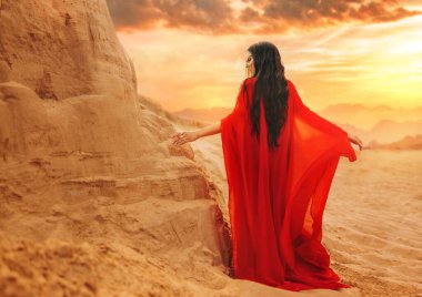 Mystery arabic woman in red long dress stands in desert long train silk fabric fly flutter in wind motion. clothes gold accessories hide face. Oriental fashion model black hair. Sand dunes sunset sun. clipart