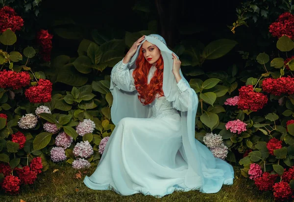 穿着白色复古服装头戴面纱的幻想女人 红头发女皇后 中世纪的公主新娘坐在花园里 绿树丛生 水草丛生 花朵繁茂 神秘形象性感的哥特式吸血鬼 — 图库照片