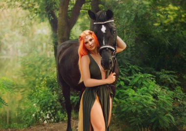 Orman perisi kız, koyu kahverengi ata dokunur ve okşar. Fantezi kadın ve at. Yeşil ağaçlar, çimenler, nehir çimenleri. Yaratıcı elf elbiseli kadın, çiçek tacıyla turuncu saçlı seksi vücut..