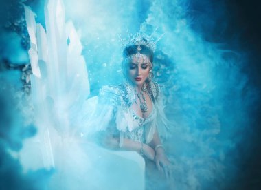 Art photo real people Fantasy woman snow queen sits on ice throne chair, white long dress train bird feathers crown icicles. Fairy tale girl frozen princess creative costume. Winter lady nature fog.