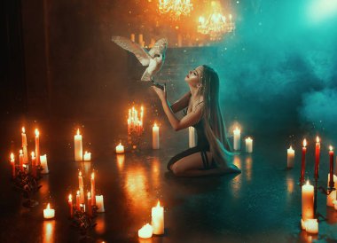 Sanat fotografı gerçek fantezi kadın gotik cadı dizlerinin üstünde oturmuş baykuş kanatlarıyla dua ediyor. Bayan Elf Sarışın, vahşi kuşları olan seksi kız. Koyu mavi duman siyah elbise ışığı 