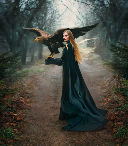 art portrait real people fantasy woman holding white-tailed eagle bird flaping open wings on hand. Elf blonde hair girl with brown bird walking in forest. Lady queen back, hunter black cape dark trees