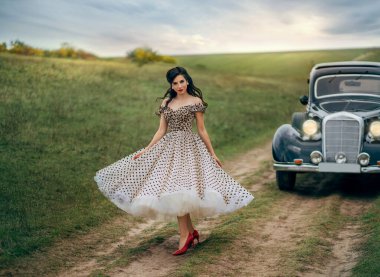 Klasik beyaz benekli, siyah retro elbisesi içinde, 50 'lerin tarzı zarif bir bayan. Pin-up saç stili Neşeli kız yolda dans ederek yürüyor. Sonbahar doğası yeşil alan gökyüzü.