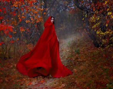 Sanatsal gotik fantezi kadın, kırmızı başlıklı kız gibi karanlık sonbahar ormanlarında yürüyor. Uzun ipek pelerin kanat çırpar, rüzgar kumaşında sallanan sinekler hareket eder. Kız prenses arkasına bakıyor. Düşen portakal, kara ağaç yolu bırakır.