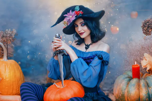Cadılar Bayramı 'nda elinde bıçakla elinde turuncu balkabağı tutan mutlu bir cadı. Şenlikli parlak sonbahar arka planı mavi dumanı hasat ediyor. Sivri uçlu şapka, mor karnaval klasik kostüm giyen kız. sihirli ışık.