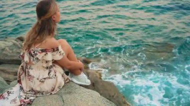 Gürültülü video. Arkaya bakan seksi genç kadın öfkeli mavi denizin taştan sahilinde oturuyor, dalgalar kayalara çarpıyor. Mutlu kız rüya görüyor, yazın tadını çıkar mavi su Costa Brava plajı İspanya